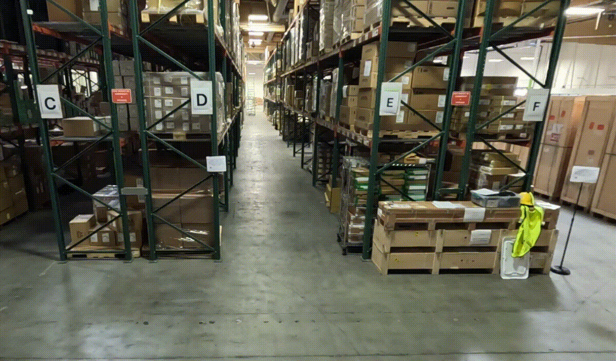 Warehouse video where a worker enters the scene and accidentally drops a box. You can also see multiple aisles and shelves in the background.